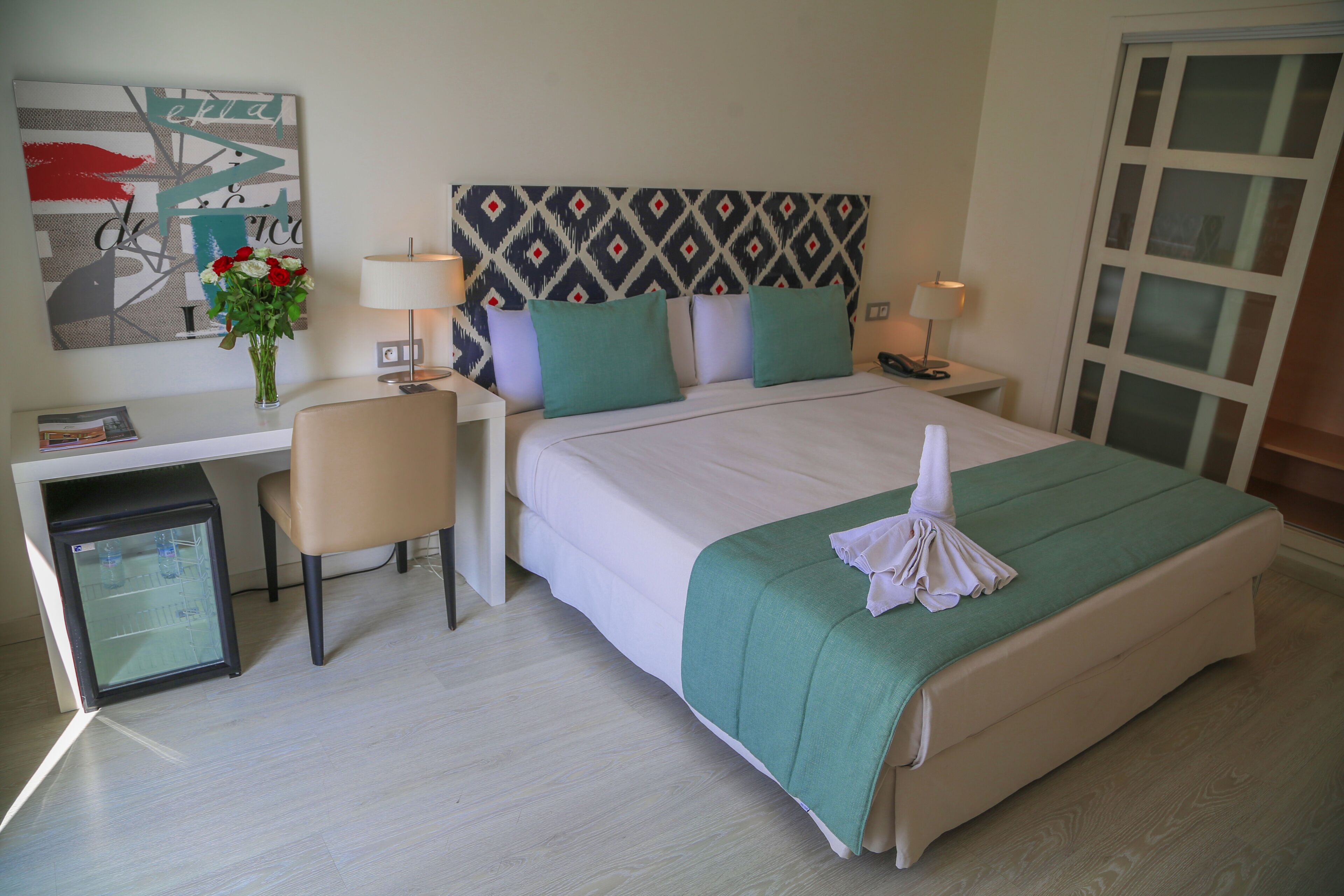 A hotel room with a bed, a desk, a chair, a refrigerator, and a wall with a geometric pattern.