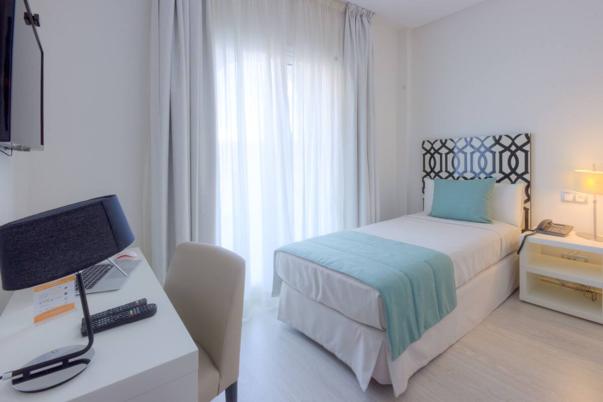 Una habitación de hotel con cama, escritorio con computadora, silla, mesita de noche con lámpara y ventana con cortinas.