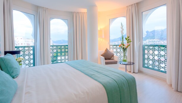 A hotel room with a large bed, a chair, a table, and a balcony with a view of the city. - Hôtel al Mandari (Tetouan)