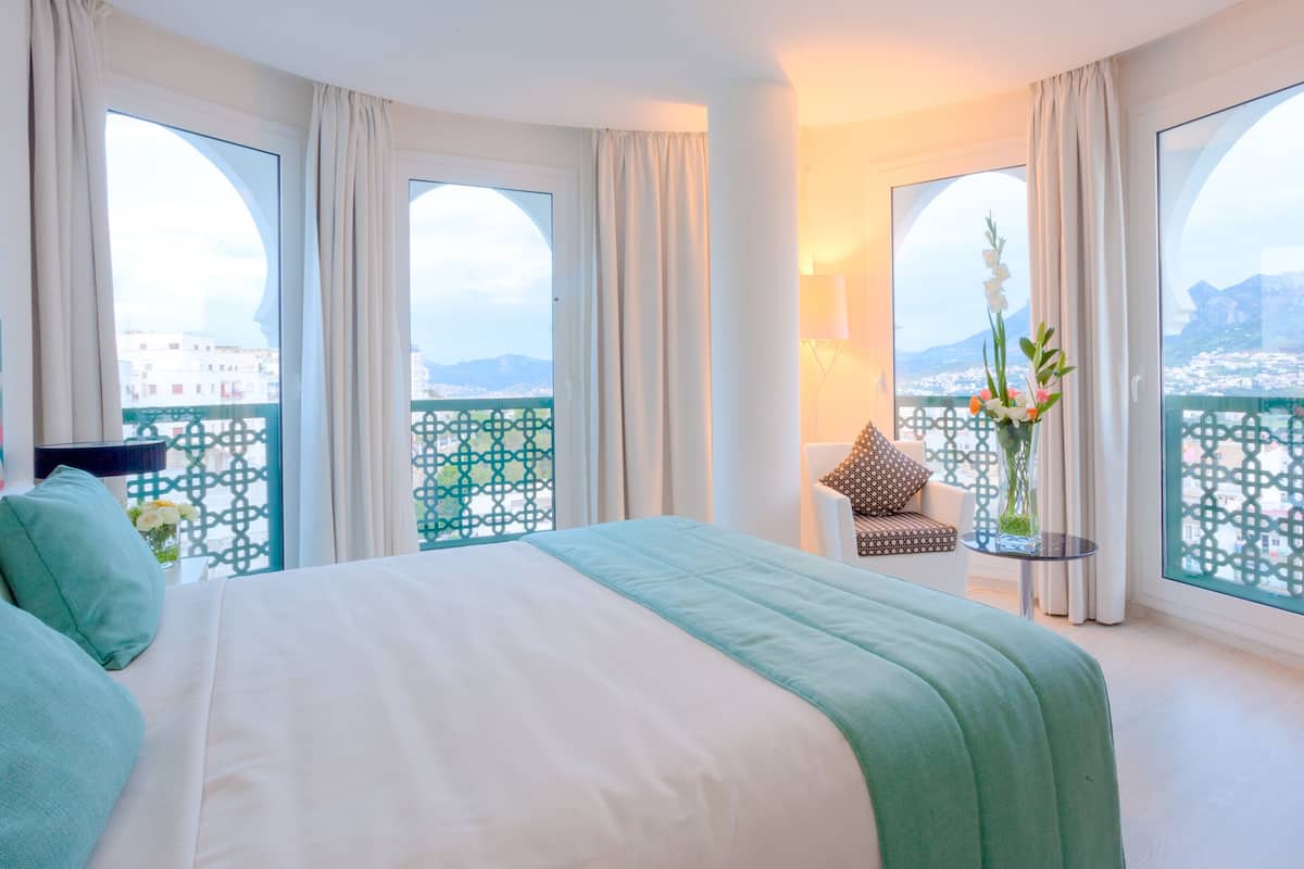 Habitación de hotel con una cama grande, una silla, una mesa y un balcón con vistas a la ciudad.