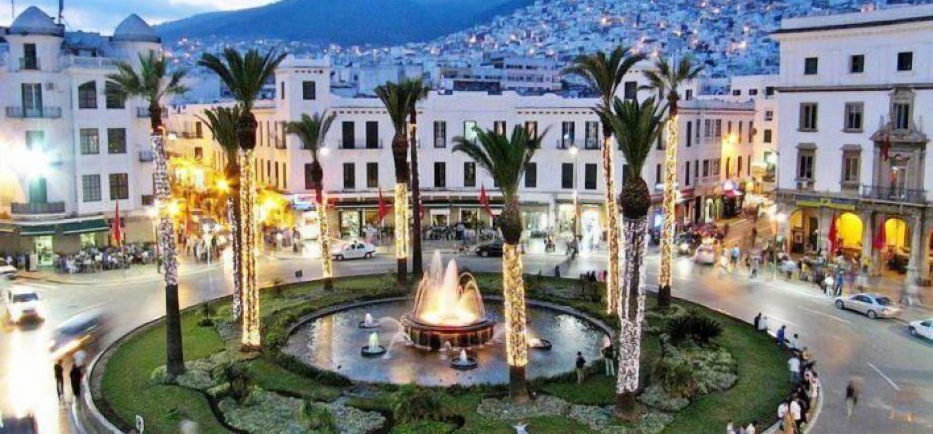 Una plaza pública con una fuente central, rodeada de palmeras y edificios de arquitectura clásica.