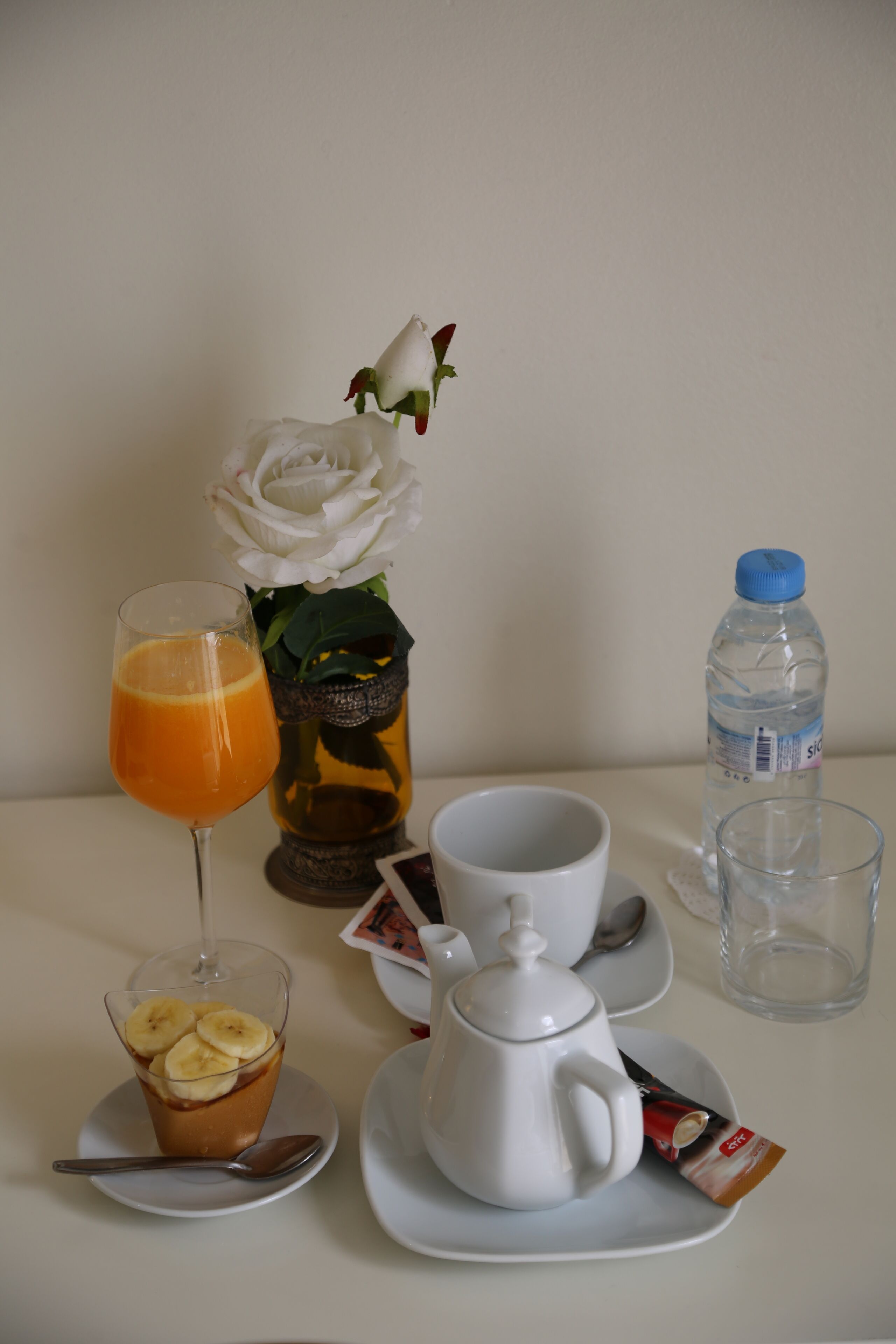 Ett bord med ett glas apelsinjuice, en kopp kaffe, en tekanna, en förpackning med grädde, en flaska vatten, en sked, en liten tallrik med bananer och en vit ros i en vas.