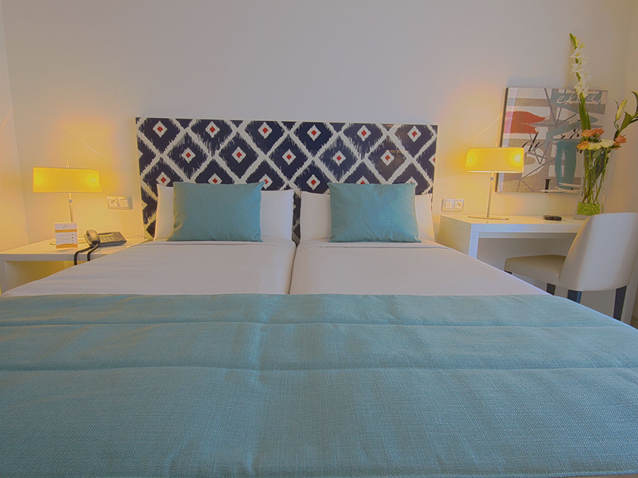 A hotel room with a large bed, two pillows, a desk, a chair, a lamp, and a vase with flowers.