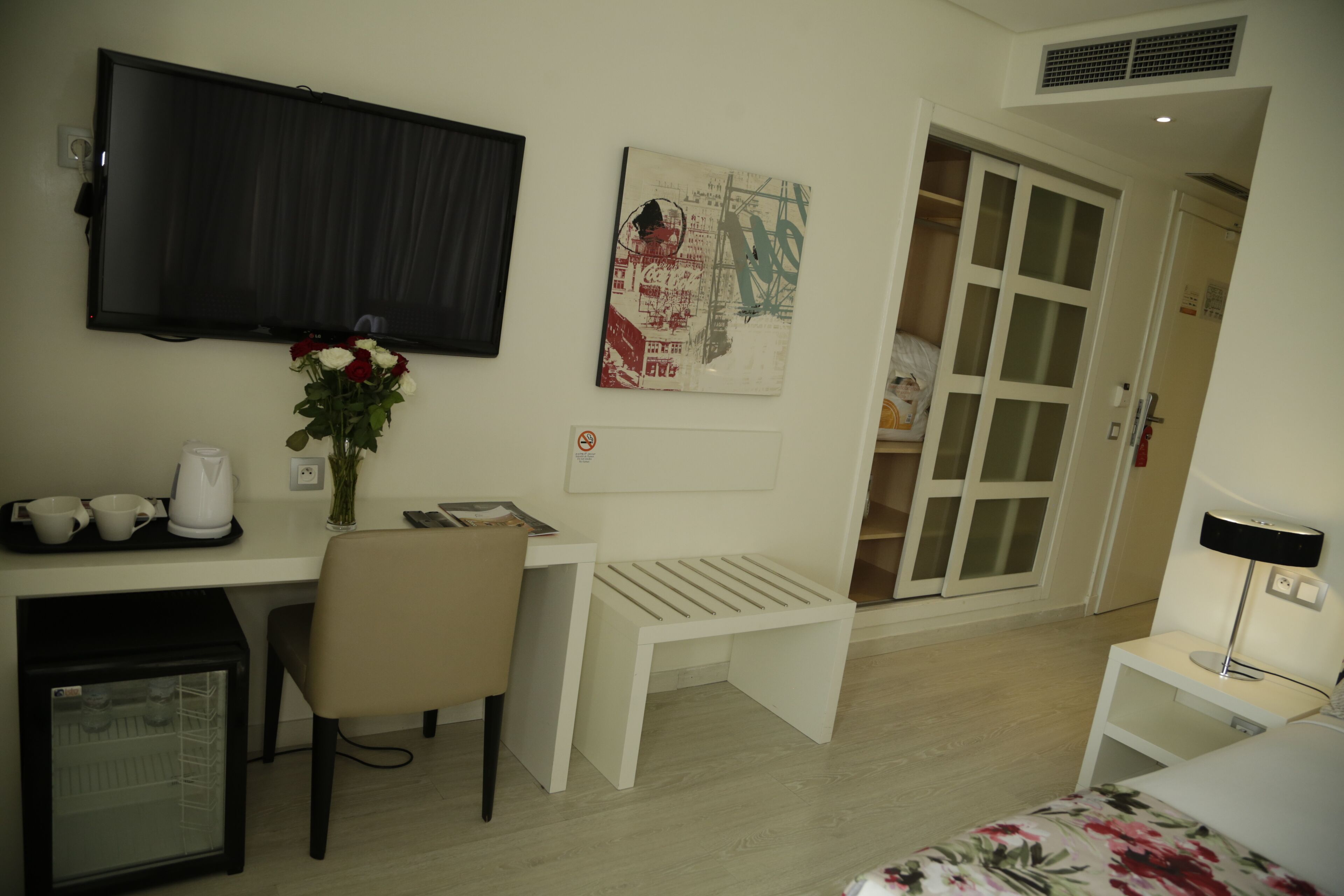 A hotel room with a bed, a desk with a chair, a television, a small refrigerator, and a bedside table with a lamp.