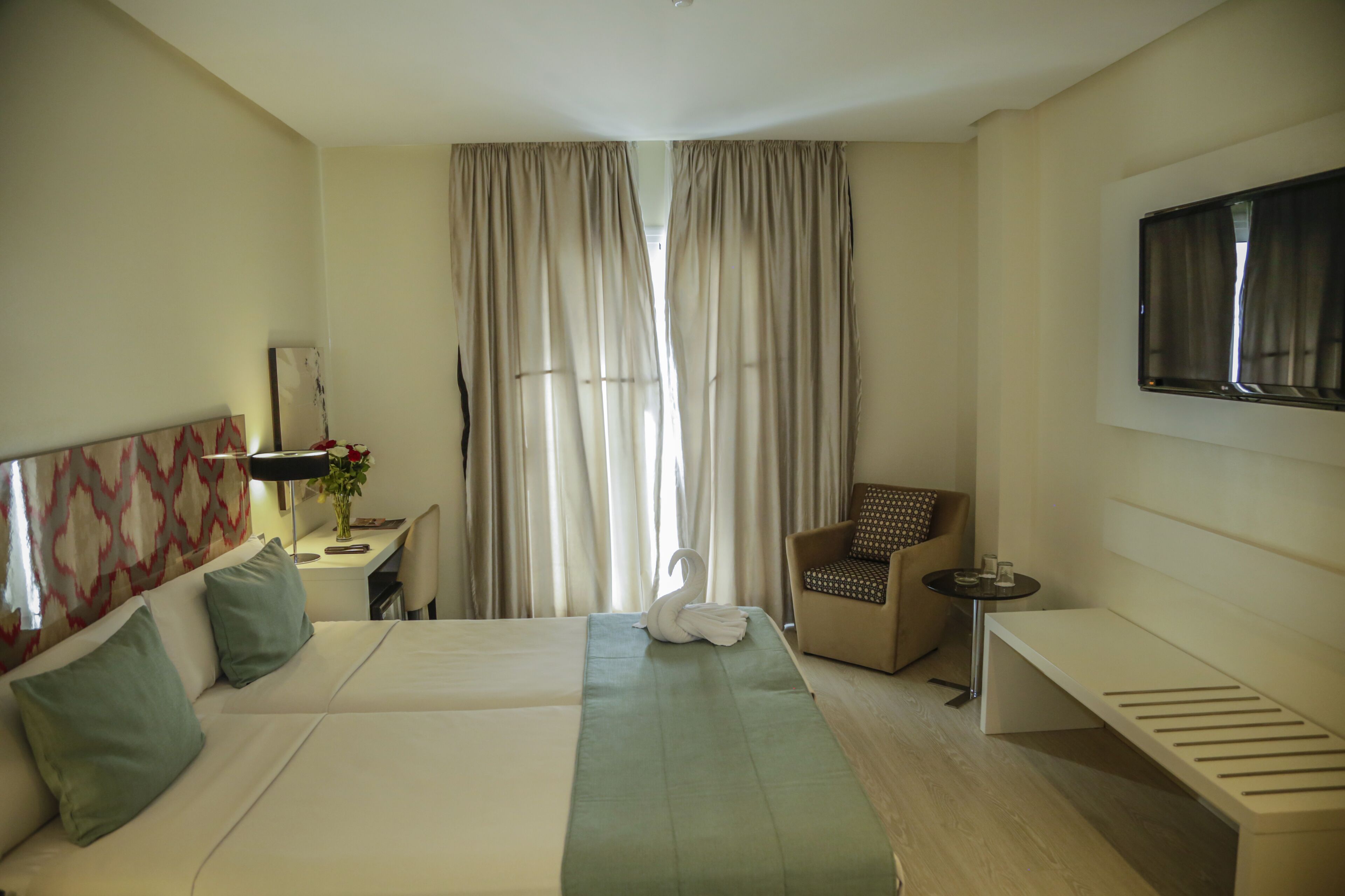 Habitación de hotel con una cama grande, un escritorio con lámpara, una silla, un banco y un televisor montado en la pared.