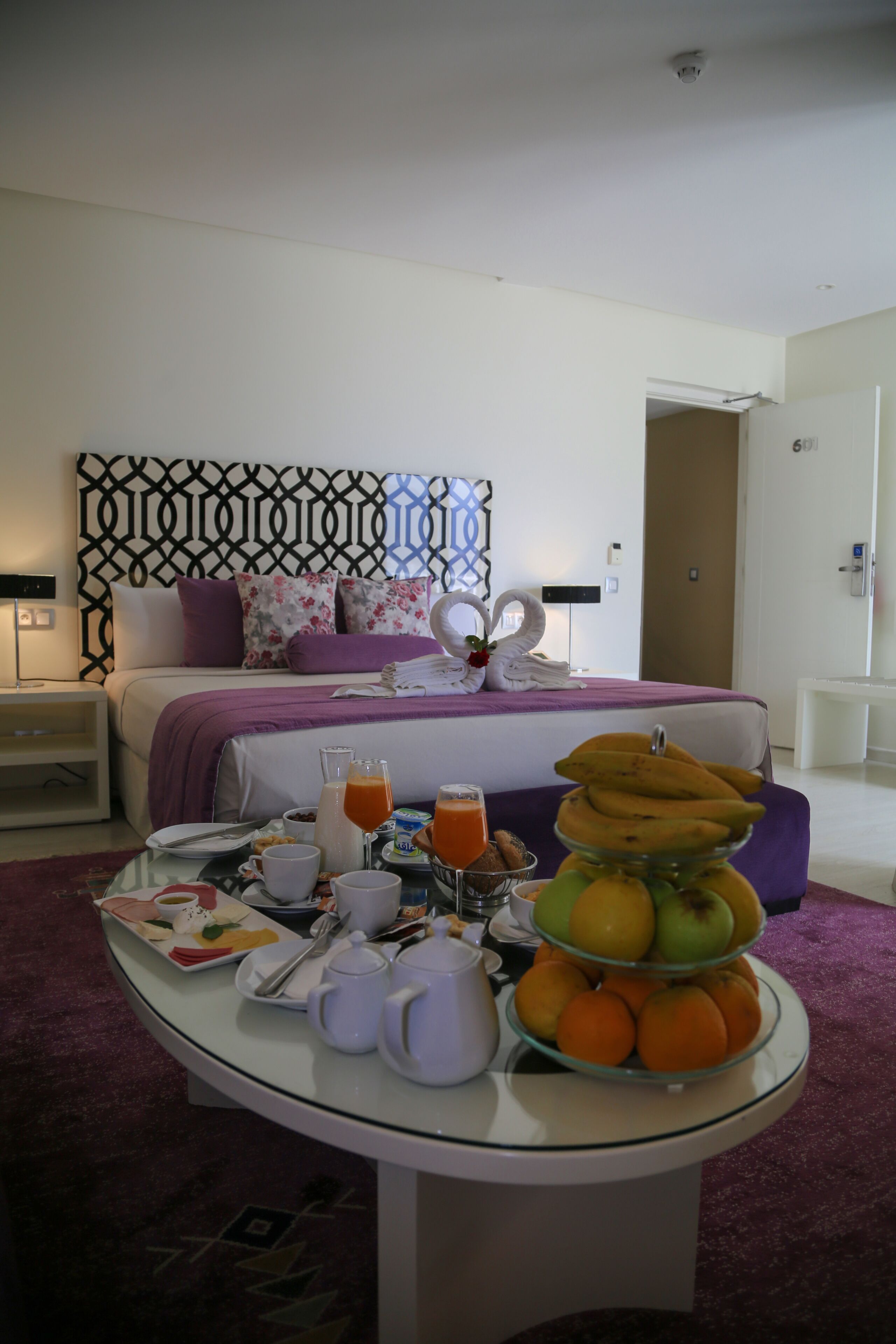 A hotel room with a neatly made bed, a breakfast table with fruits, a fruit bowl, and a vase of flowers.