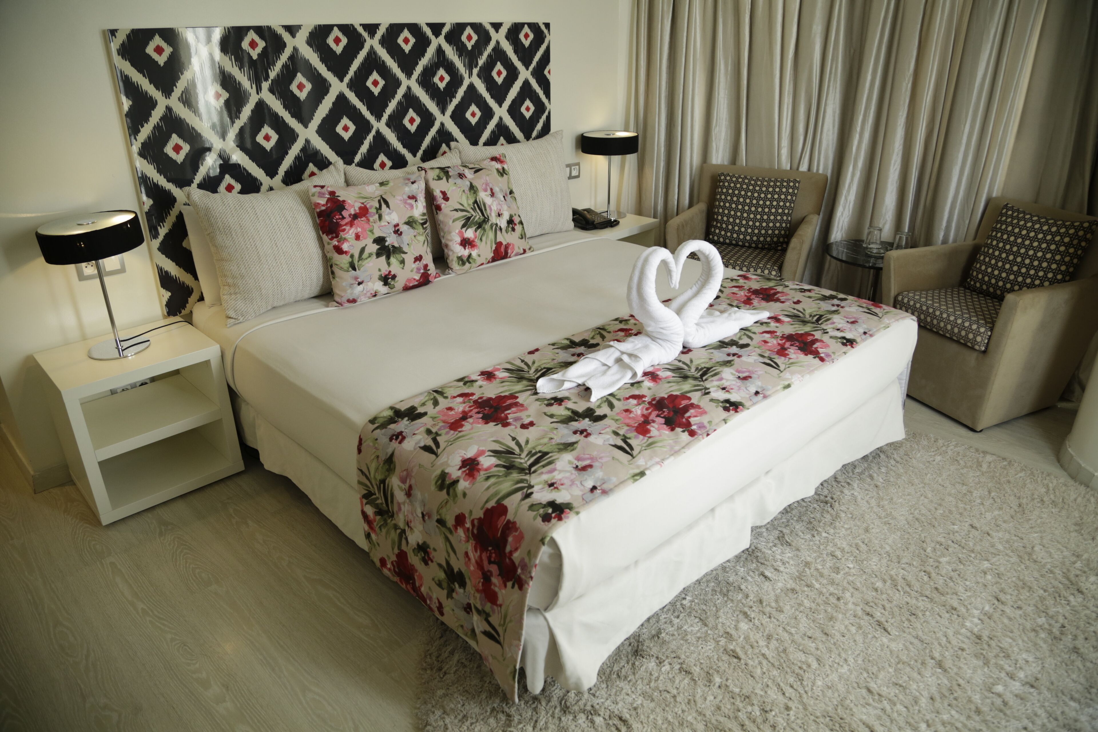 A hotel room with a large bed, floral bedspread, two nightstands, a lamp, a chair, and a headboard with a geometric pattern.