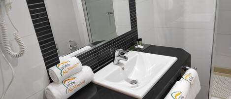 Un baño con lavamanos blanco, encimera negra y cabina de ducha con paredes de vidrio. Hay toallas con el logotipo de 'Hotel Al Mandari' y un teléfono blanco en la pared.