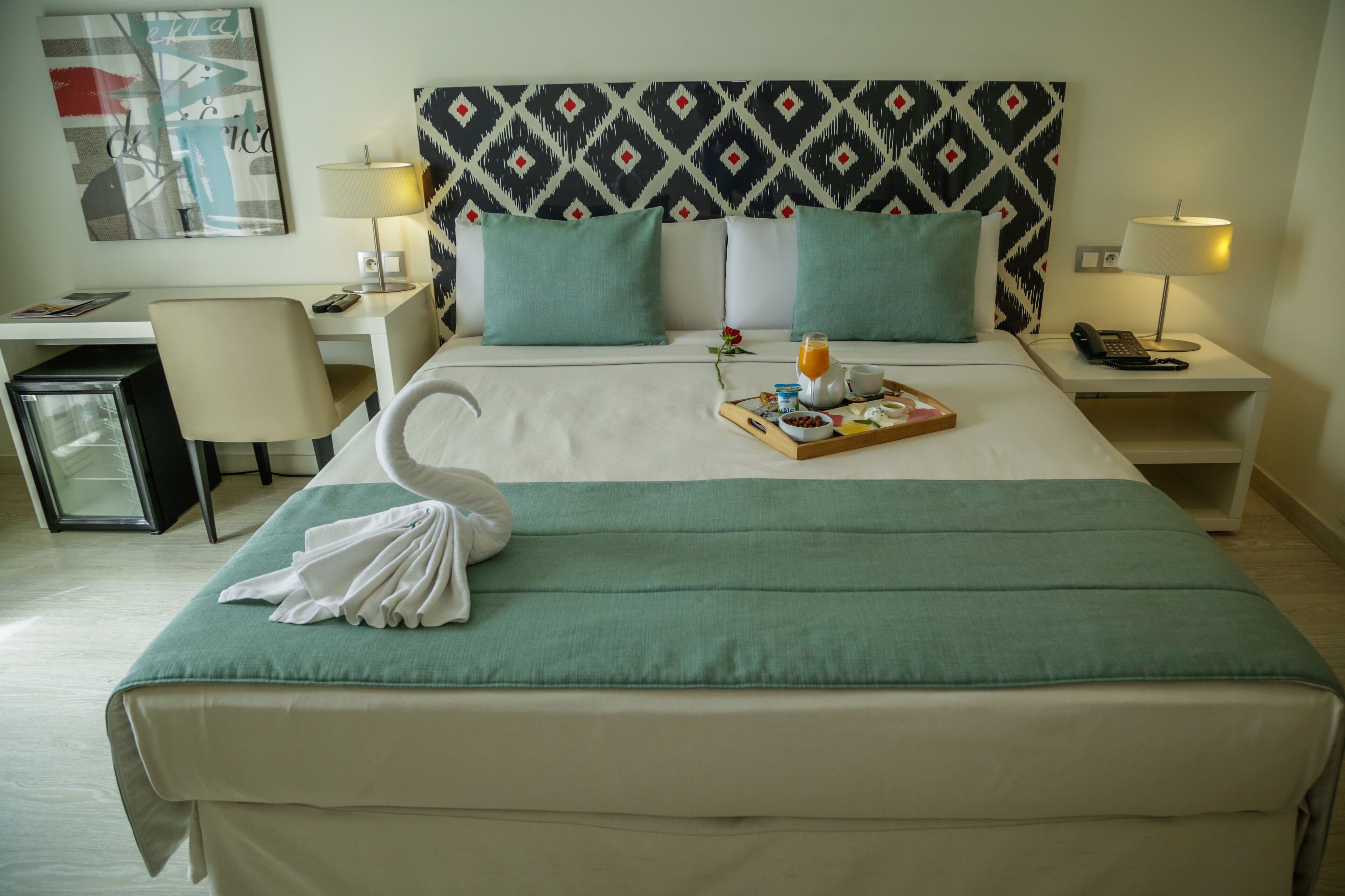 A hotel room with a bed, a desk, a chair, a nightstand, a lamp, and a small refrigerator.