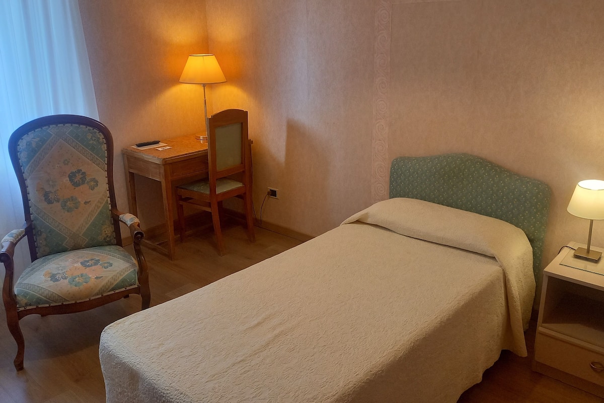 Une chambre d’hôtel avec un lit simple, un bureau avec une chaise, une lampe et une table de chevet.