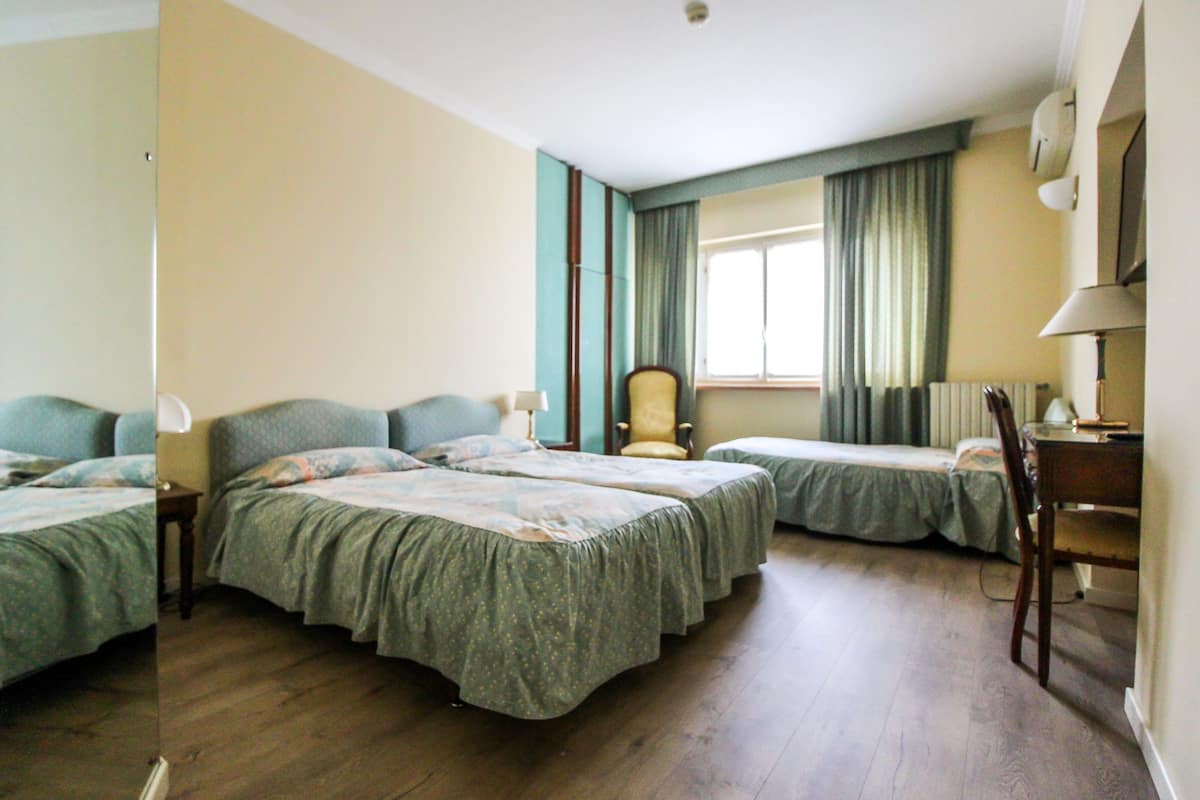 Une chambre d’hôtel avec trois lits simples, une chaise, un bureau et une fenêtre avec des rideaux.