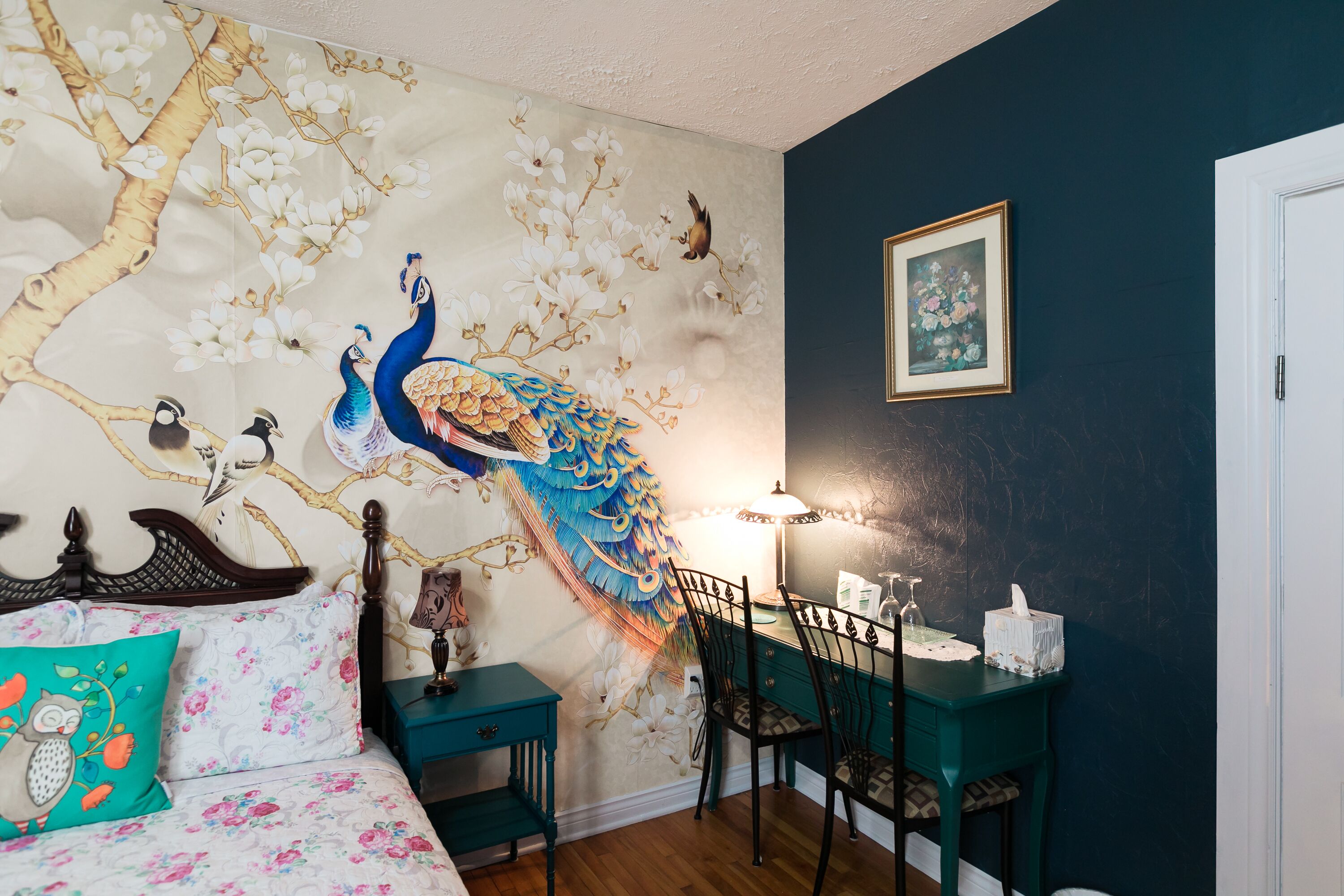 Een slaapkamer met een muurschildering van een pauw, een bed met bloemenkussens, een bureau en een stoel.