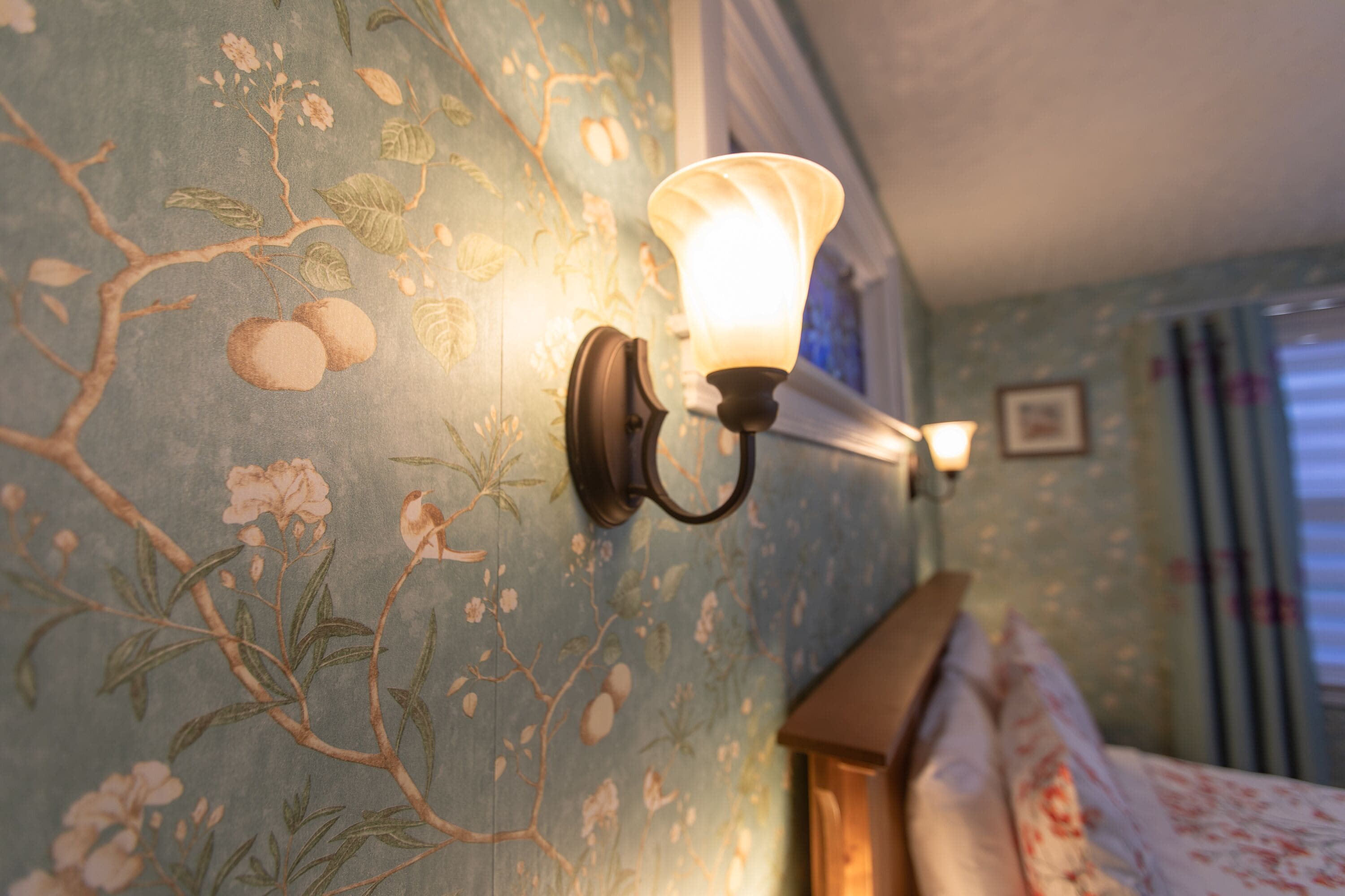 Eine Wand mit blumigem Wandpapier, eine Wandleuchte und ein Bett mit hölzernem Kopfteil.