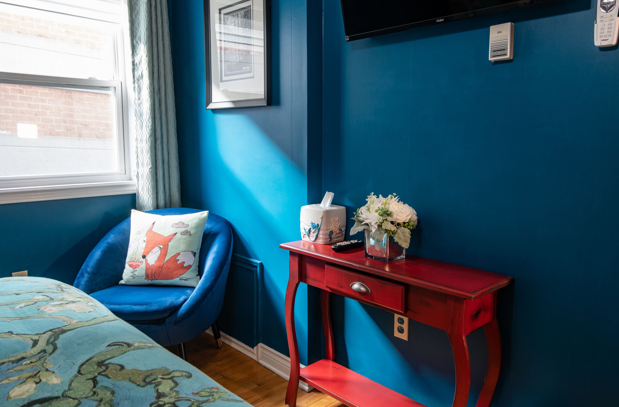 A bedroom with a blue chair, a red nightstand, a bed with a floral bedspread, and a TV mounted on the wall.