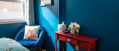 A bedroom with a blue chair, a red nightstand, a bed with a floral bedspread, and a TV mounted on the wall.