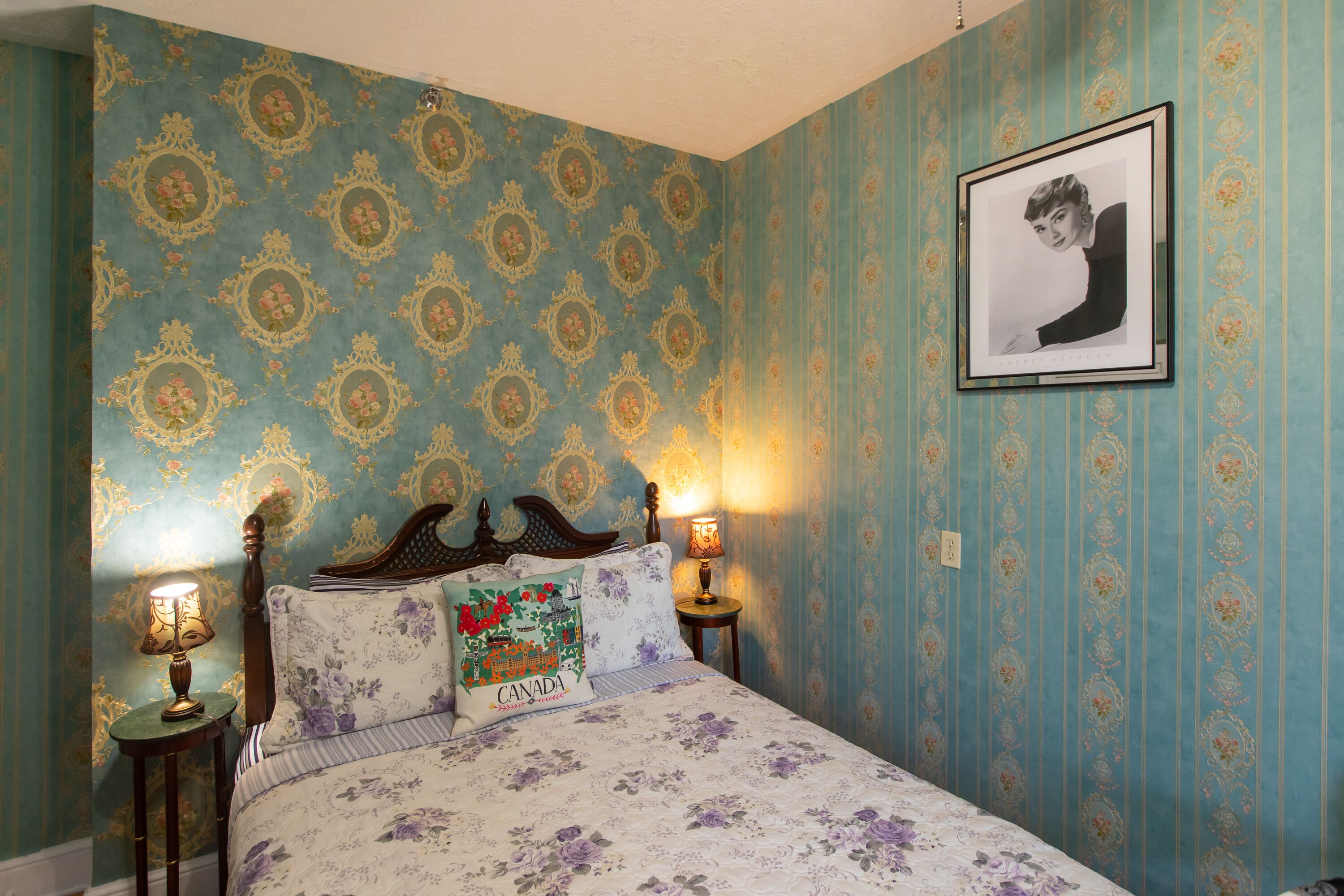 Une chambre à coucher avec un papier peint à motifs floraux, un lit avec une literie à motifs floraux, deux lampes de chevet et un tableau encadré au mur.