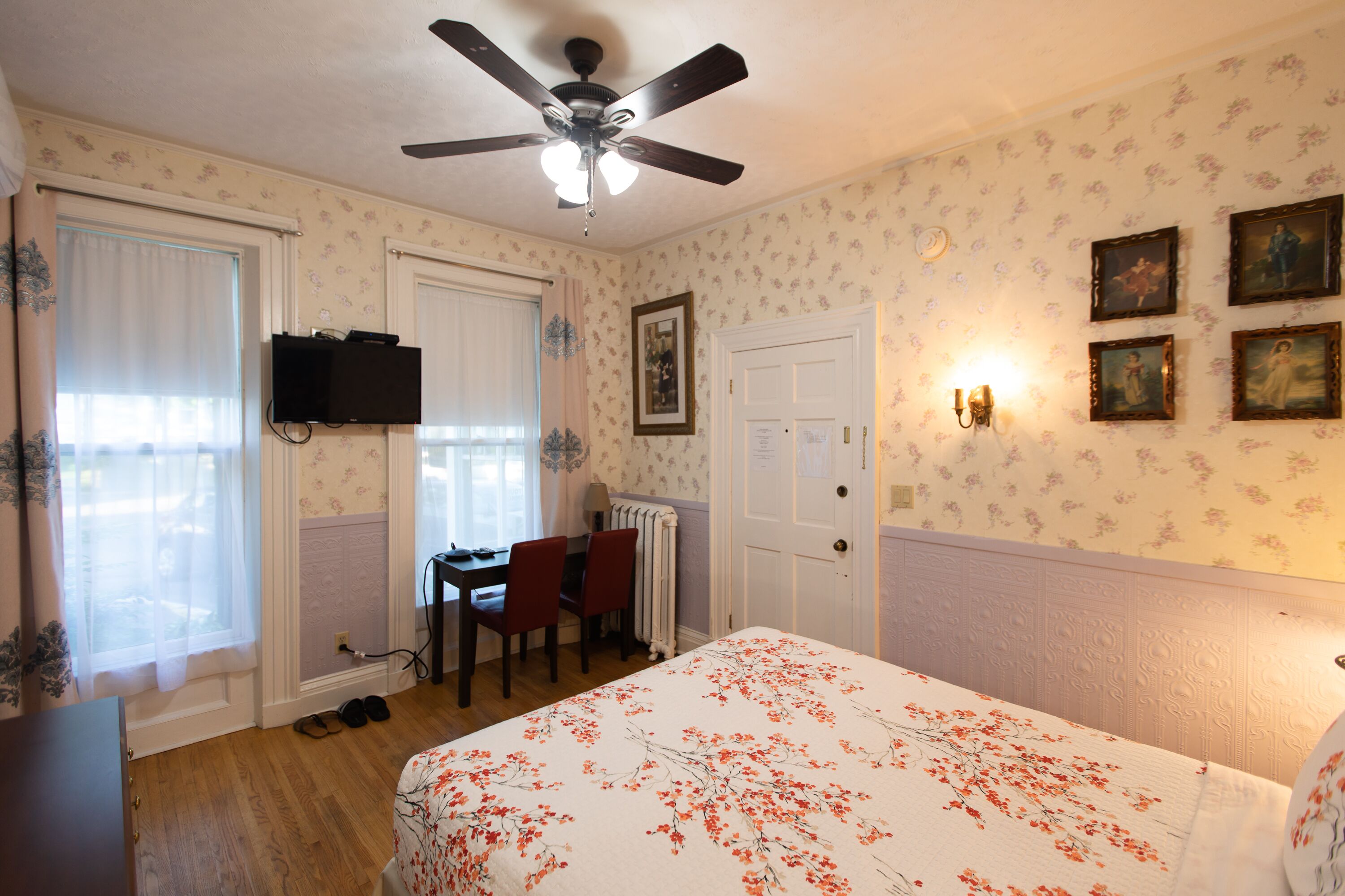 Een slaapkamer met een bed, een plafondventilator, een bureau met stoelen, een radiator en lijstjes met foto's aan de muur.