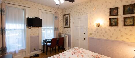 Een slaapkamer met een bed, een plafondventilator, een bureau met stoelen, een radiator en lijstjes met foto's aan de muur.