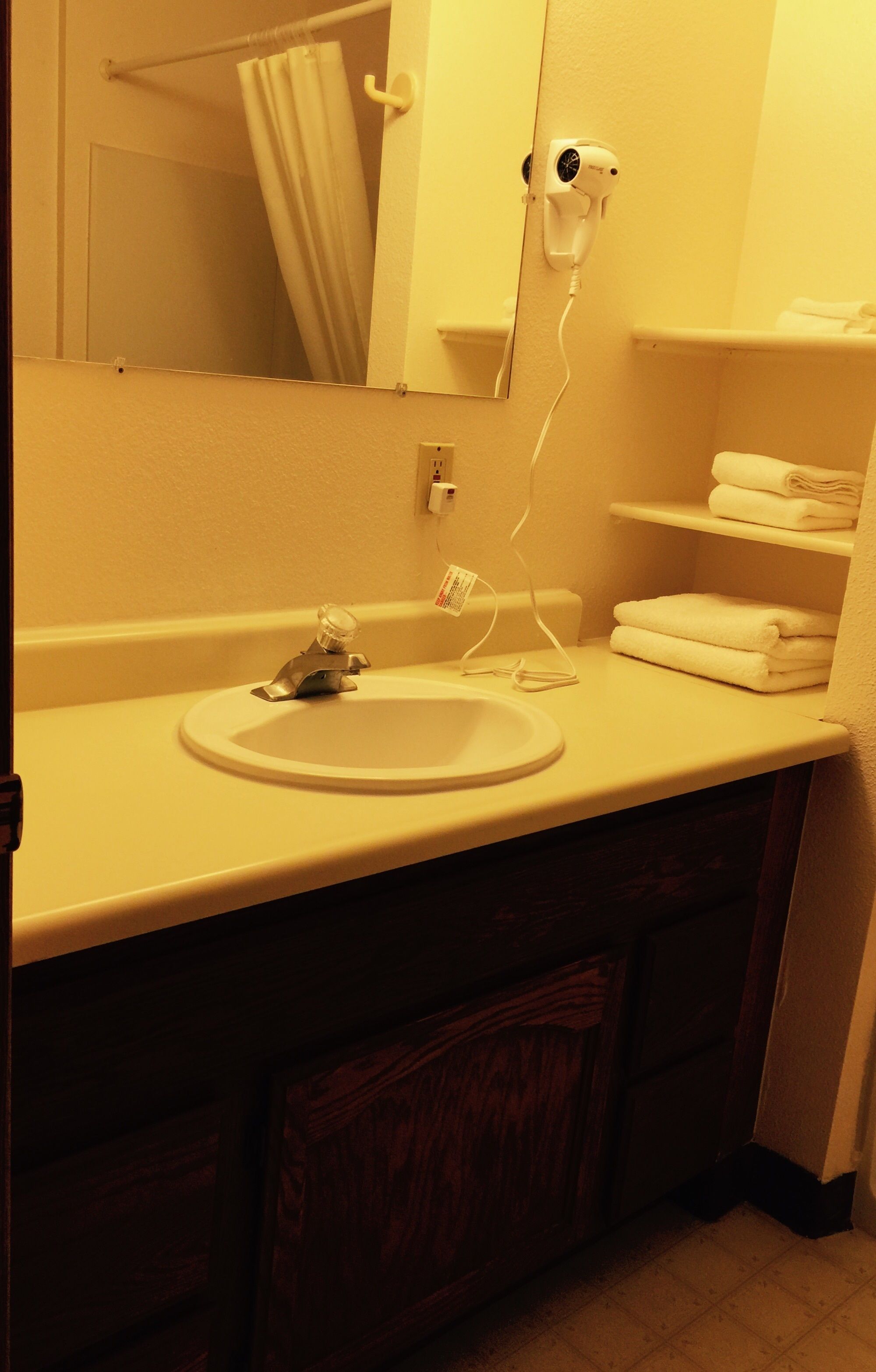 A bathroom with a white sink, a mirror, a hairdryer on the wall, and a shelf with folded towels.