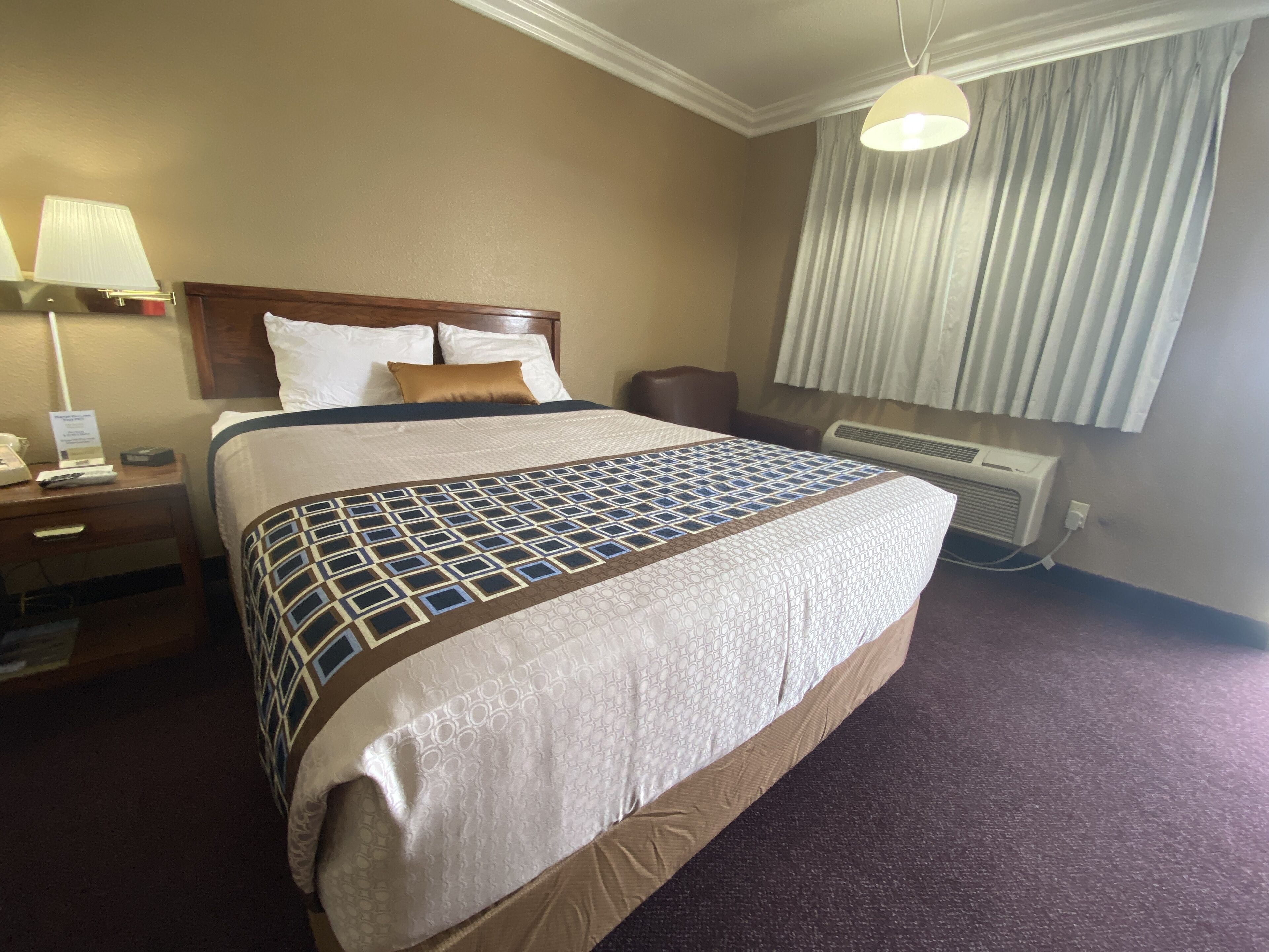 A hotel room with a large bed, a bedside table with a lamp, a chair, a window with curtains, and an air conditioning unit.