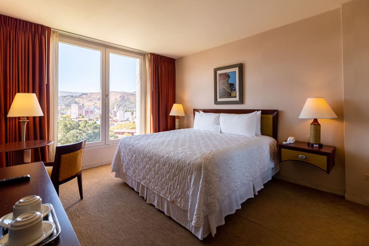 A hotel room with a large bed, two bedside tables with lamps, a chair, a desk with a remote control, and a view of the city through the window.
