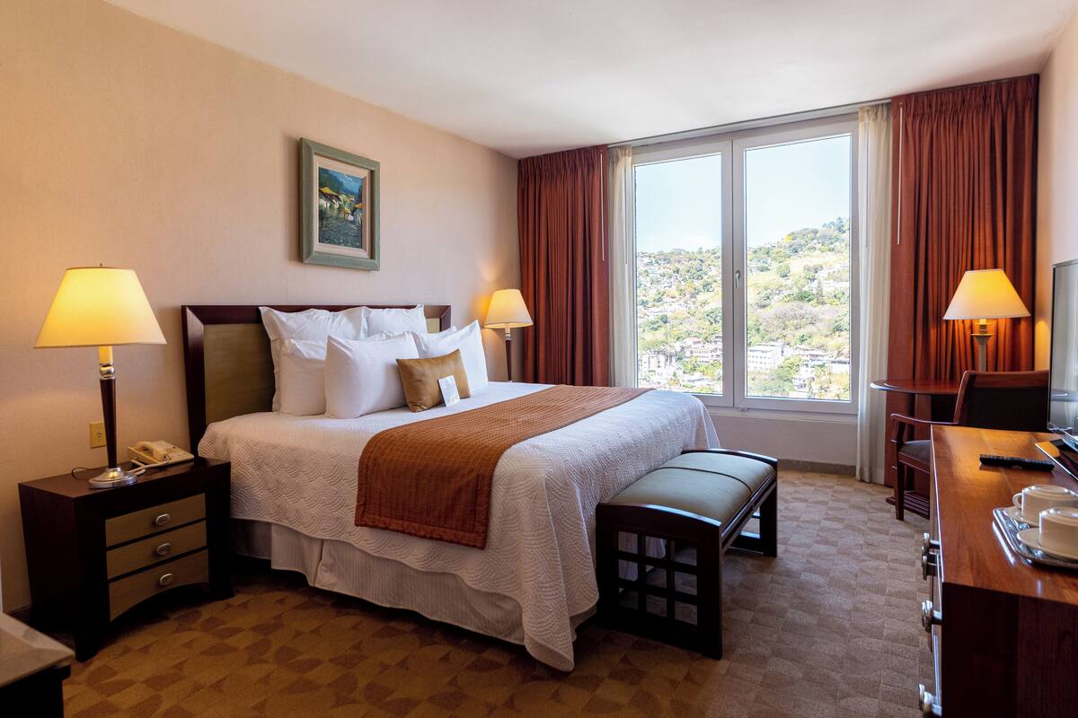 A hotel room with a large bed, two bedside tables with lamps, a desk with a chair, and a view of a hilly landscape through the window.