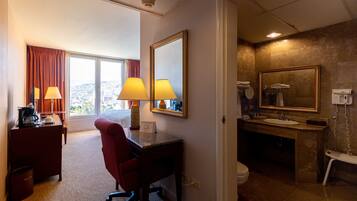 A hotel room with a desk, chair, two lamps, a large mirror, a bathroom with a sink, toilet, and a shower.