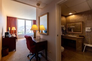 Habitación de hotel con escritorio, silla, dos lámparas, un espejo grande, y baño con lavamanos, inodoro y ducha.