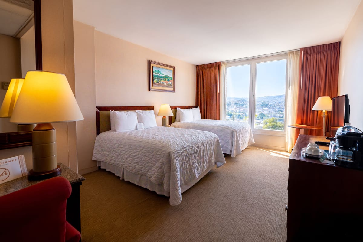 A hotel room with two beds, a large window with curtains, a painting on the wall, and a desk with a lamp and a coffee maker.