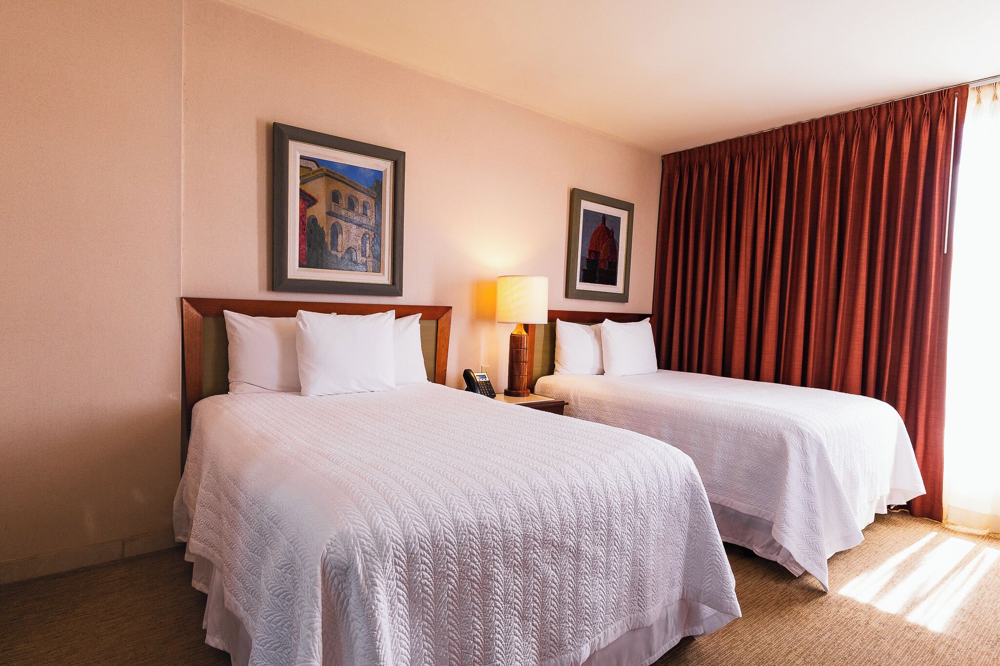 A hotel room with two beds, a lamp, a telephone, and framed pictures on the wall.