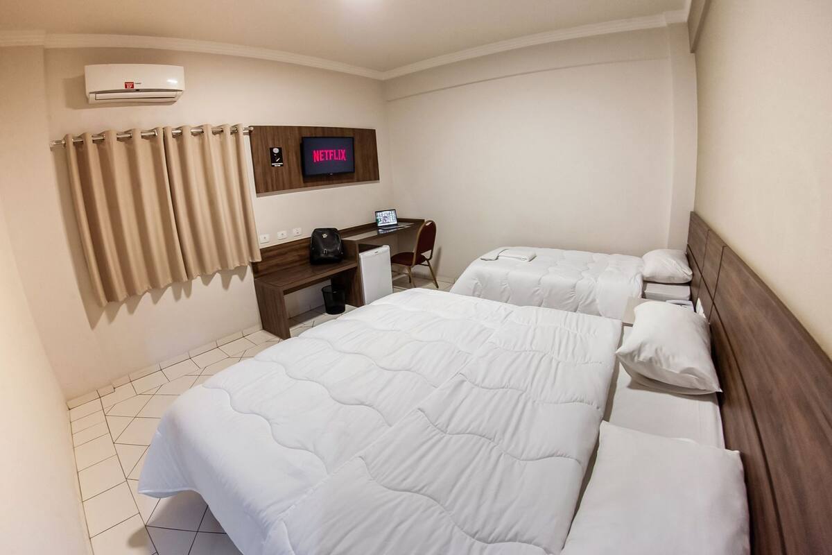 Ein Hotelzimmer mit zwei Betten, einem Schreibtisch mit einem Computer, einem Fernseher, der 'NETFLIX' anzeigt, und einer Klimaanlage.