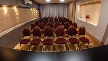 Un salón de conferencias con filas de sillas rojas, un ventanal con cortinas y un equipo de aire acondicionado instalado en la pared.