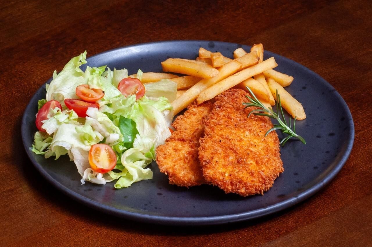 Een bord met een gepaneerd biefstukje, friet en een salade.