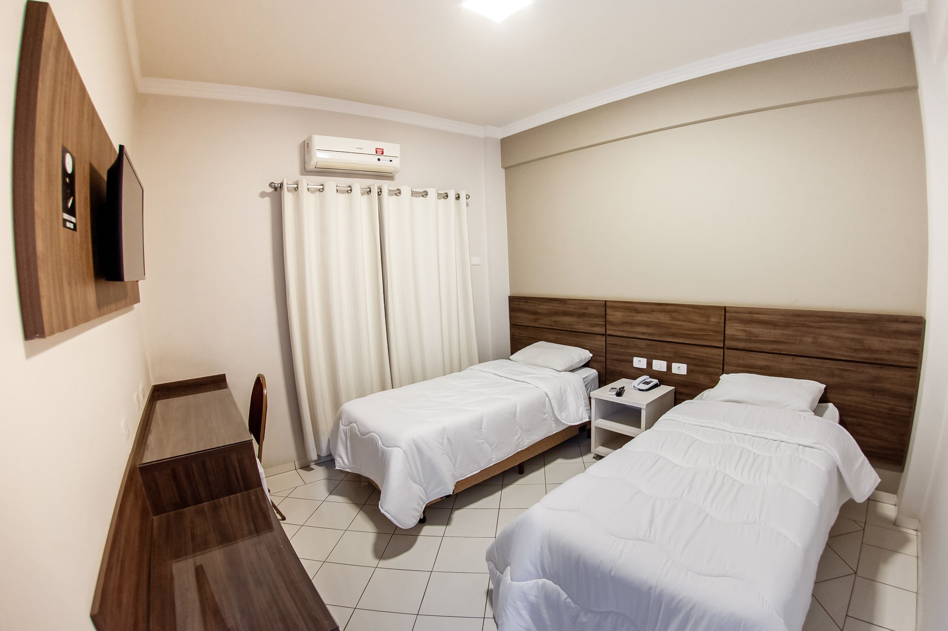 Een hotelkamer met twee eenpersoonsbedden, een bureau, een stoel, een aan de muur bevestigde televisie en een airconditioning.