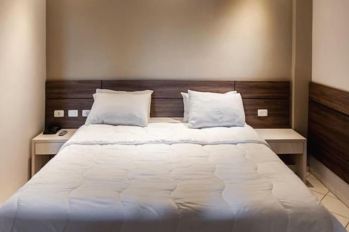 Una cama bien hecha con edredón blanco y almohadas, flanqueada por dos mesitas de noche con lámparas.