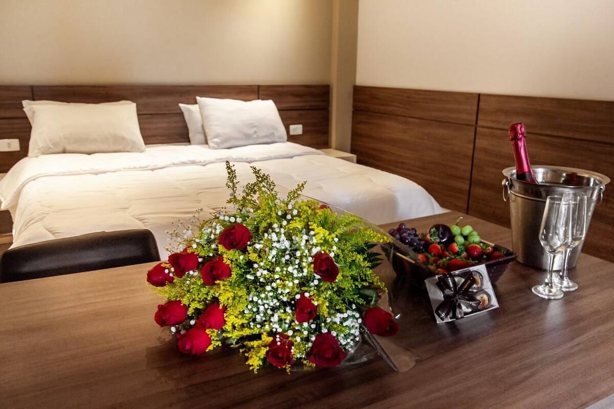 Ein Hotelzimmer mit einem ordentlich bezogenem Bett, einem Holfnachtkästchen mit einem Strauß roter Rosen, einer Obstschale und einer Champagnerflasche.
