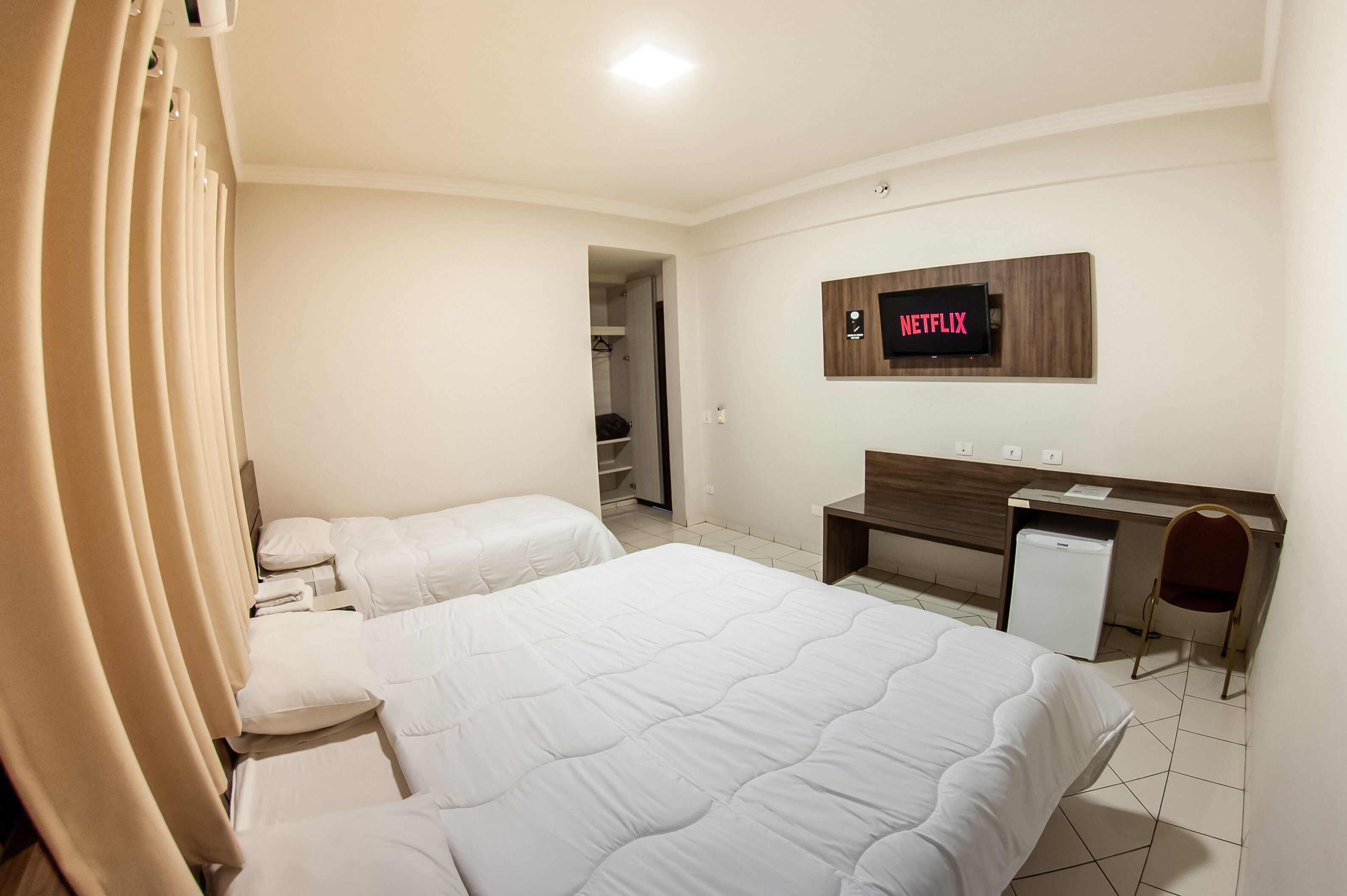 Une chambre d’hôtel avec deux lits, un bureau, une chaise, un téléviseur fixé au mur et un petit réfrigérateur.