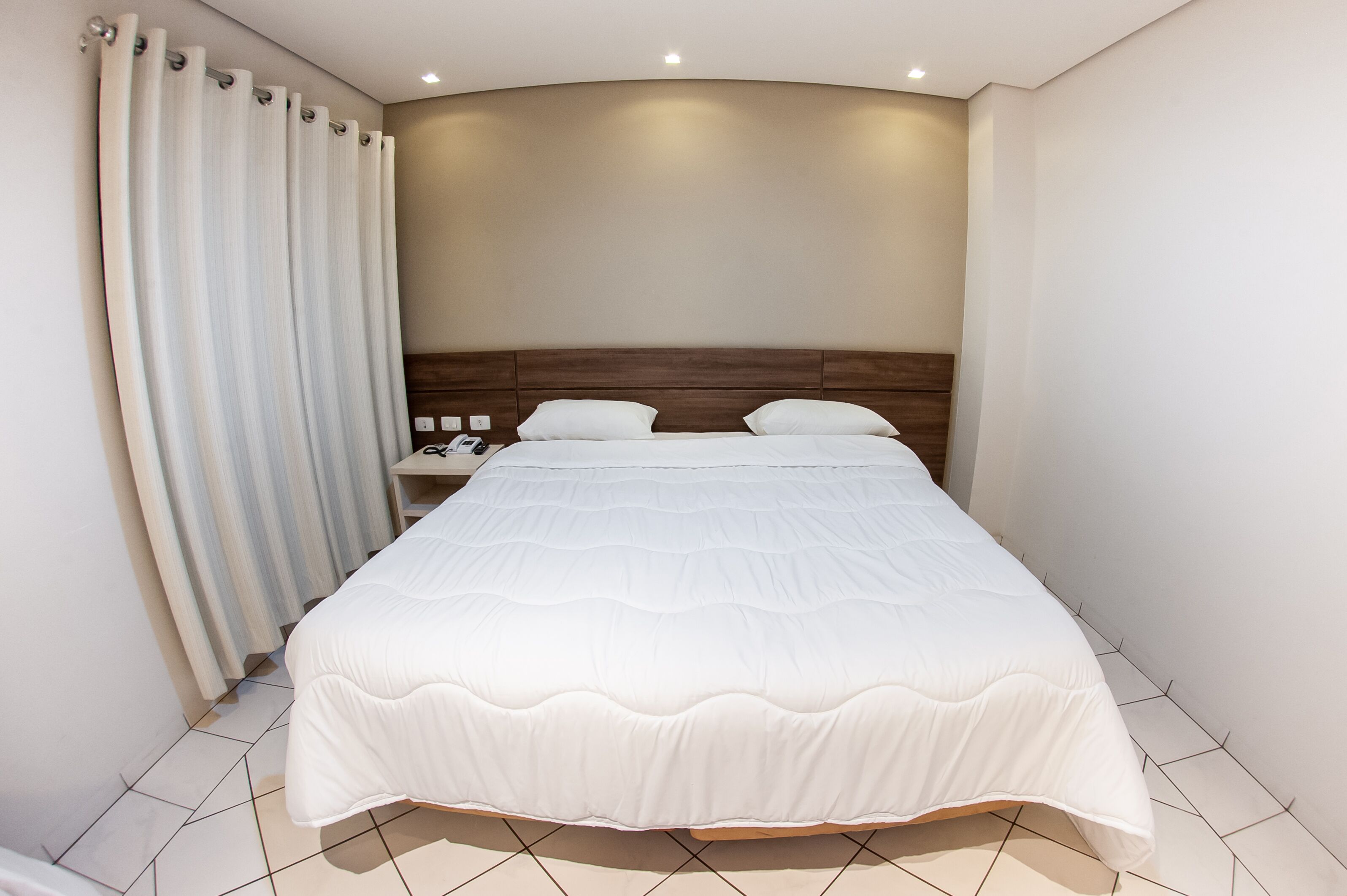 A hotel room with a large bed, a wooden headboard, a nightstand with a phone, and a curtain.