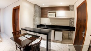 A compact kitchen with a microwave, sink, and a small refrigerator. There is a dining table with chairs and a wooden door on the left. - London Hotel (Londrina)