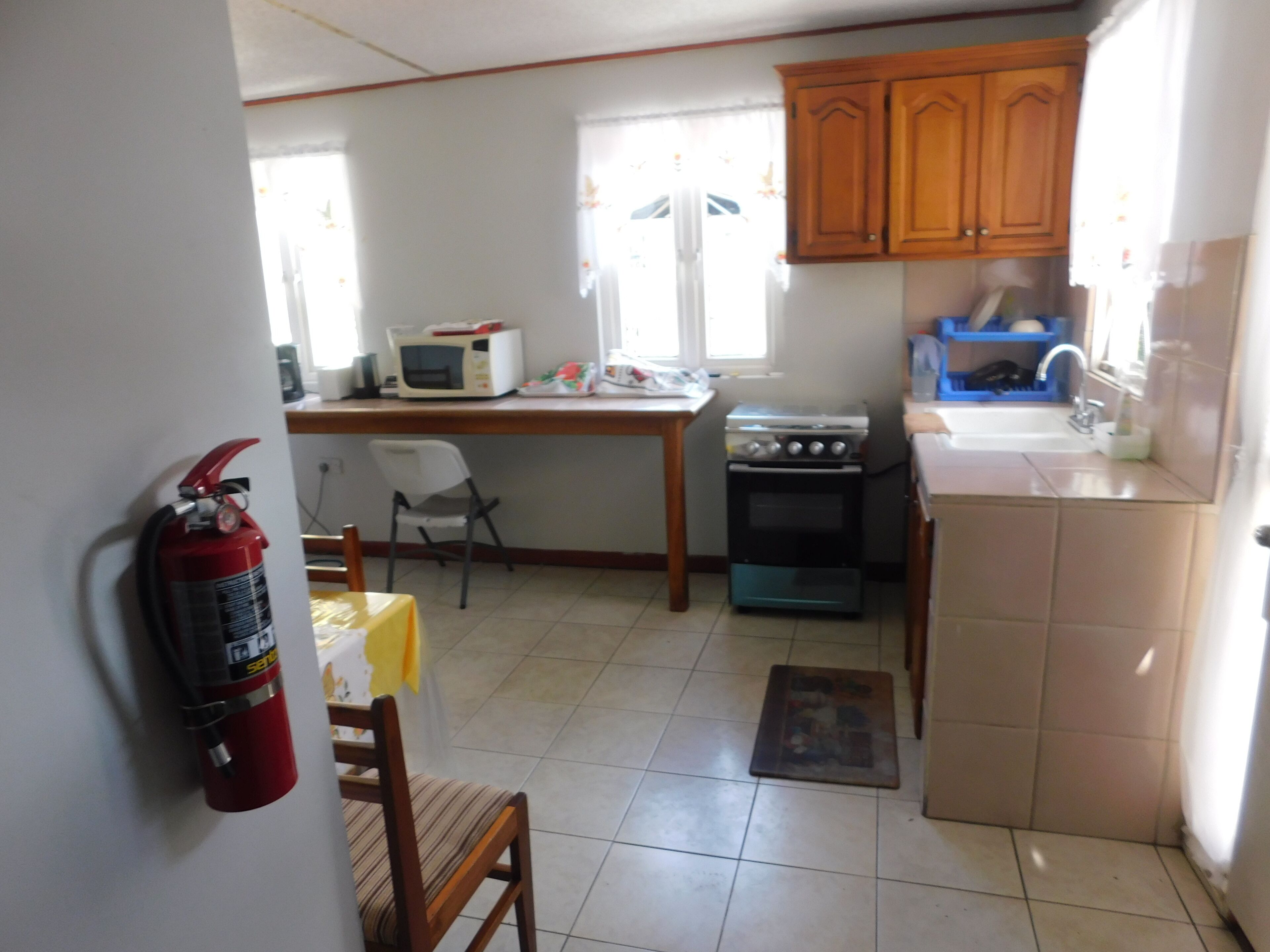 Uma cozinha compacta com micro-ondas, fogão e pia. Há um extintor de incêndio na parede e uma cadeira perto da mesa.