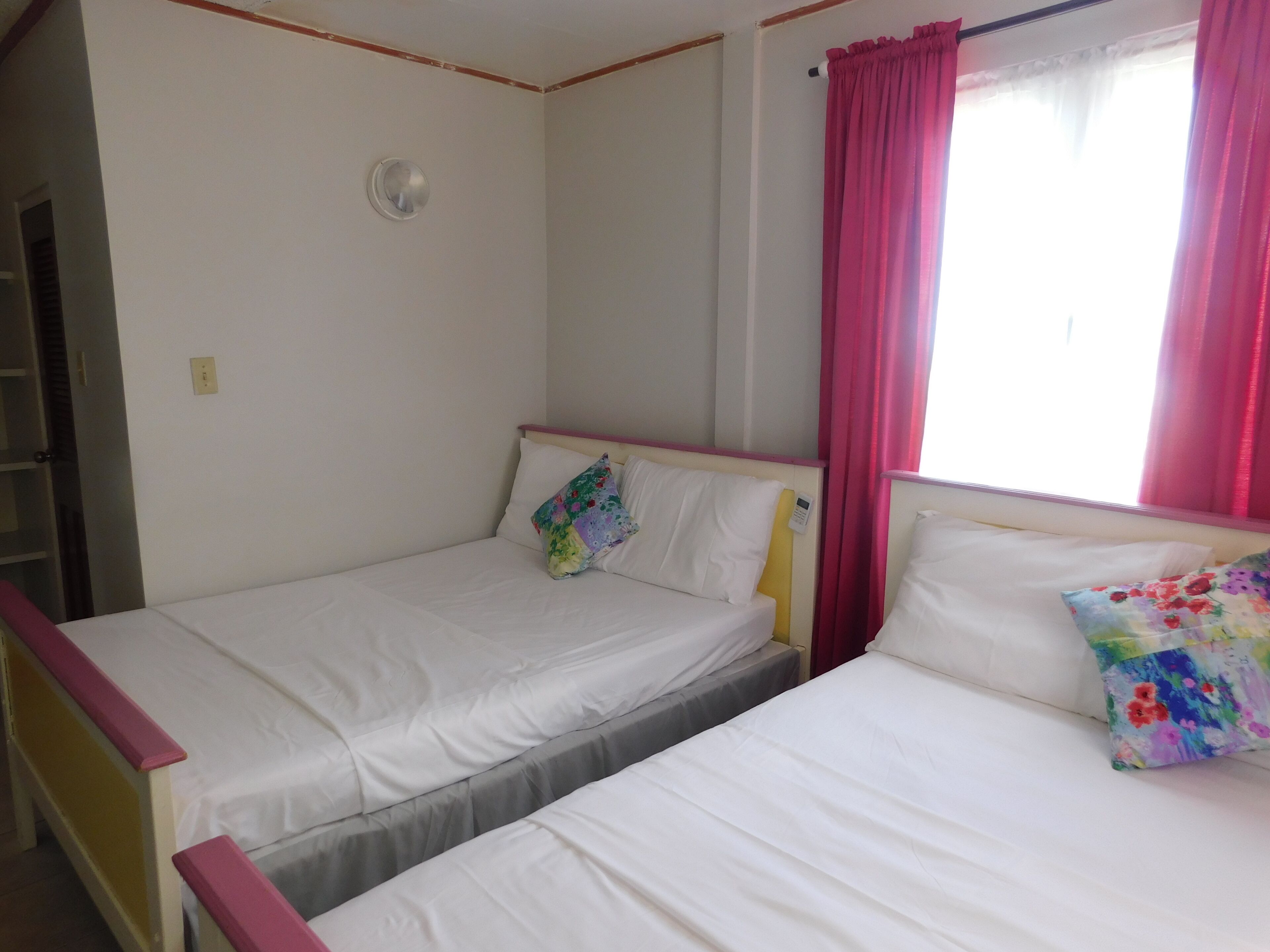 Two single beds with white bedding, a red curtain, and a small window with pink curtains.