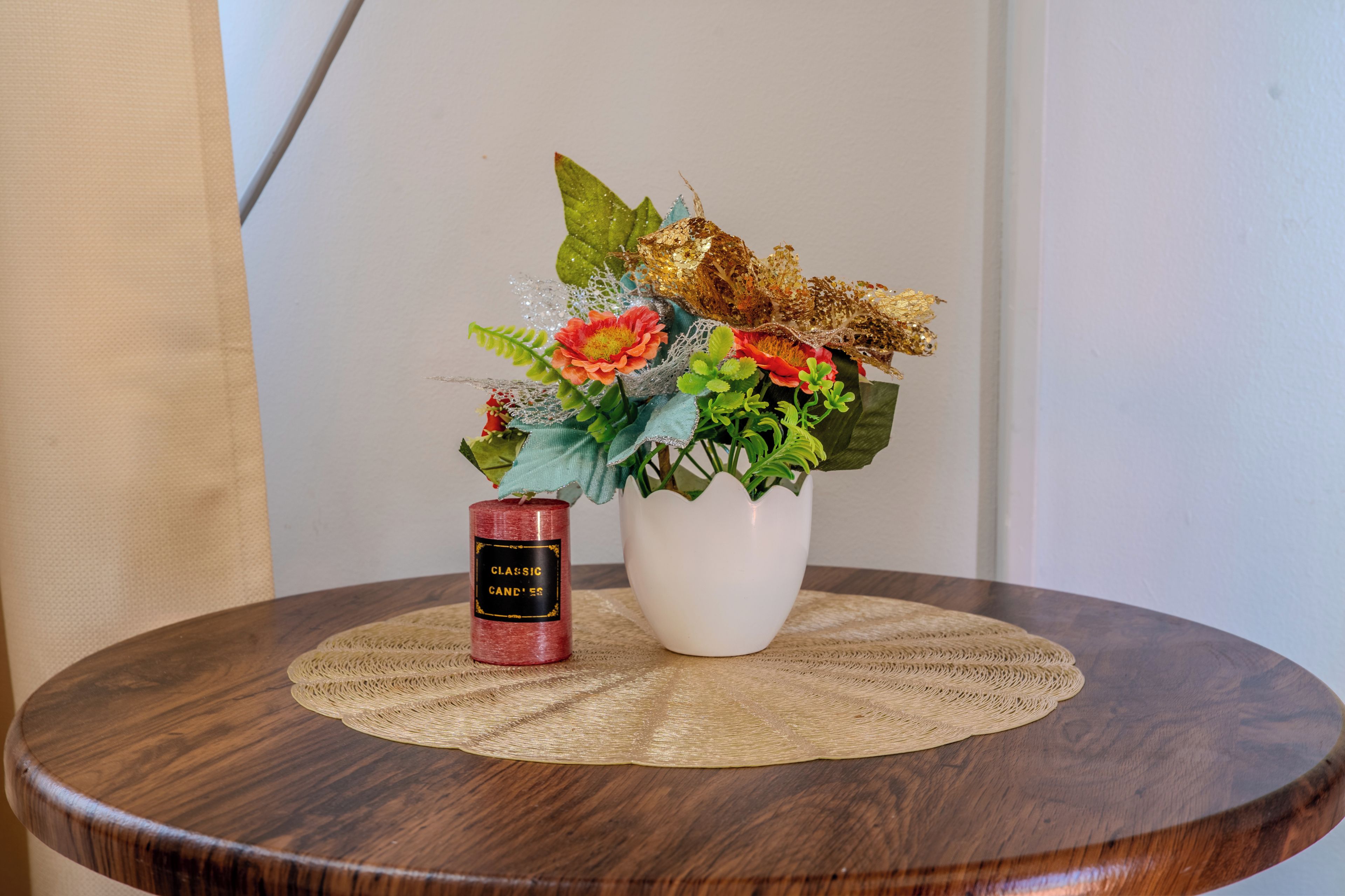 Une table ronde en bois surmontée d’un set de table en fibres tressées, un vase blanc contenant des fleurs artificielles et une bougie rouge portant l’étiquette « BOUGIES CLASSIQUES ».