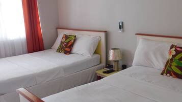 Een hotelkamer met twee enkele bedden, elk met een witte spreidek en een kleurrijk kussen. Daartussen staat een nachtkastje met een lamp en een afstandsbediening.