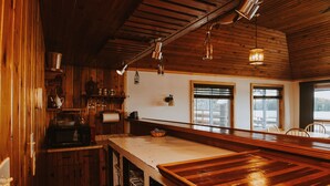 A rustic kitchen with wooden cabinets, a large wooden counter, hanging pendant lights, and a view through the windows. - Lang Lake Resort (Unorganized North Sudbury)