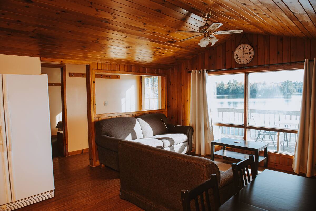 A cabin interior with a wooden ceiling, a ceiling fan, a sofa, a dining table, and a view of a lake through large windows.