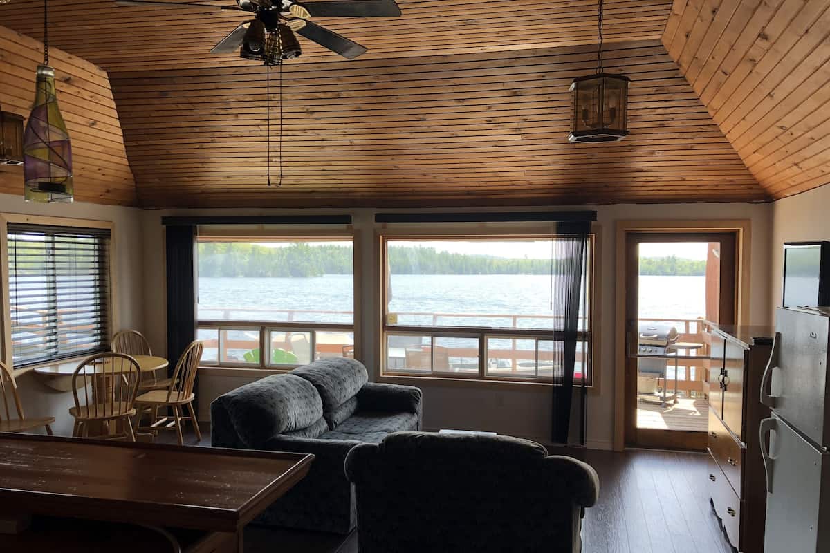 A cozy room with a wooden ceiling, a sofa, a dining table, and large windows overlooking a lake.