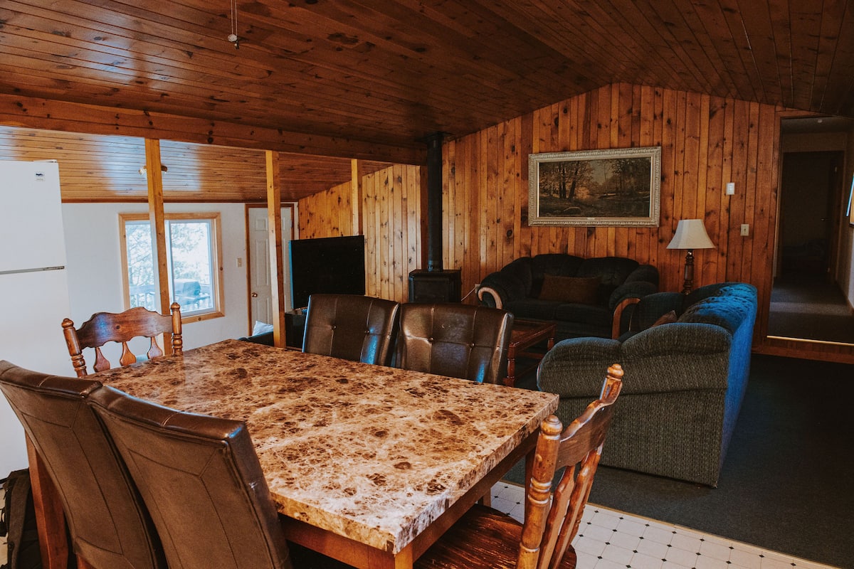 A wooden-paneled room with a dining table, chairs, a sofa, and a television.