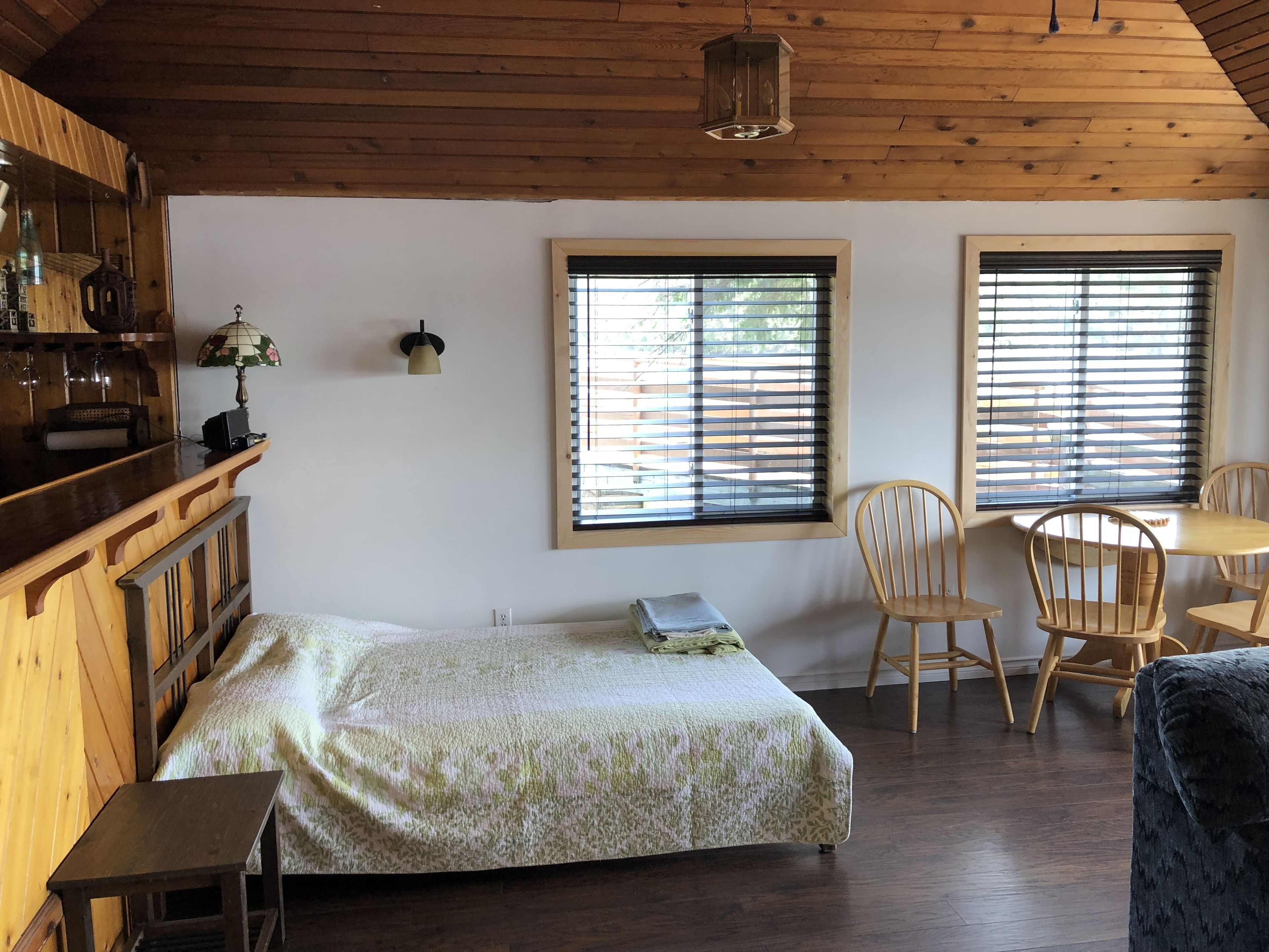 Una habitación acogedora con una cama, una pequeña mesa y un rincón comedor con sillas. Hay ventanas con persianas, una lámpara colgante y techo de madera.