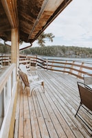 Terraza de madera con sillas y una mesa, con vista a un cuerpo de agua y zona boscosa.