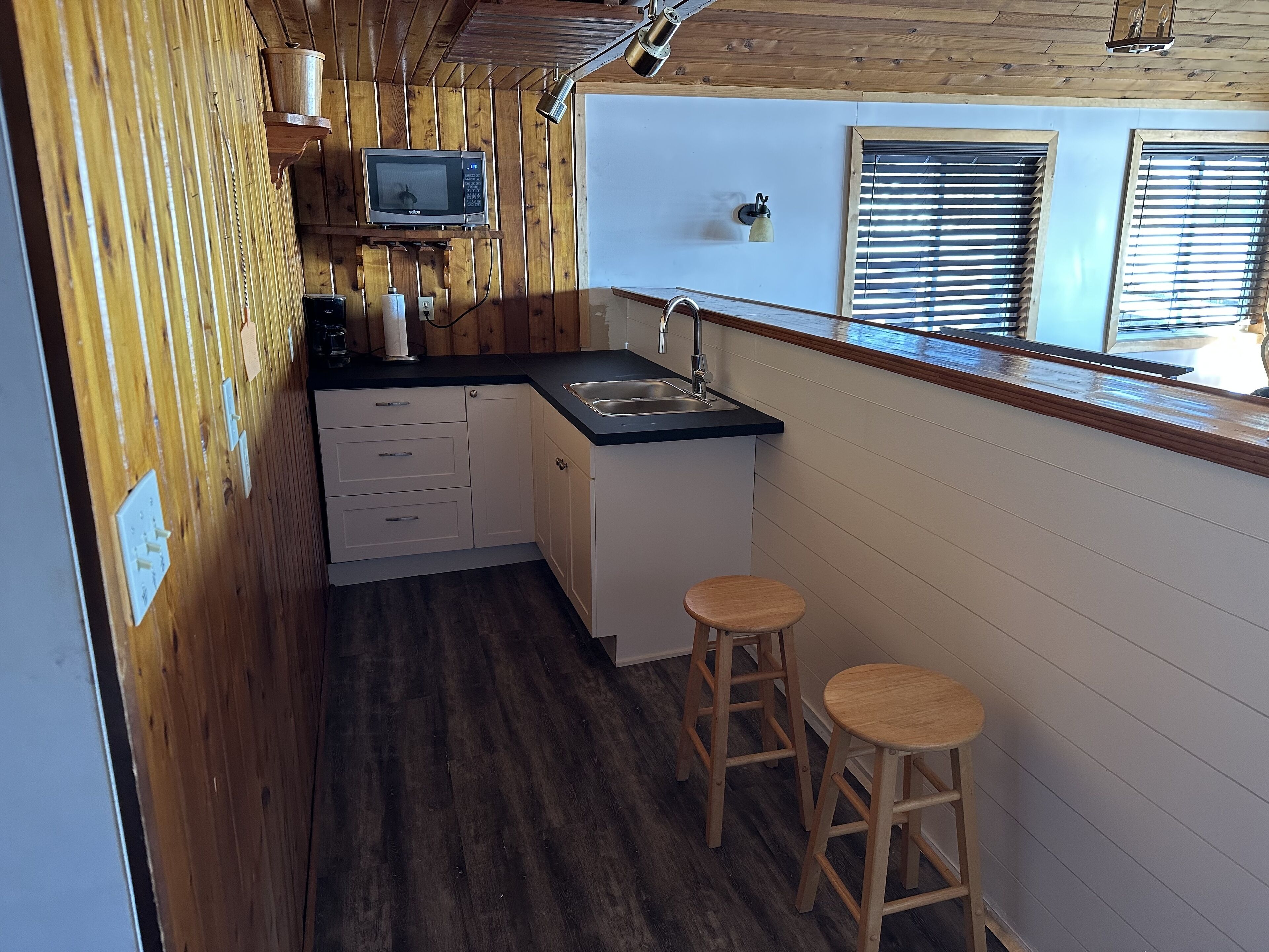 A compact kitchen with wooden walls, a microwave, a sink, and two stools.